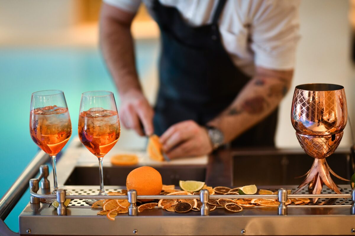 Glasses of orange colored drink, male bartender slicing oranges